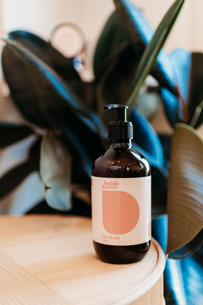 Freddie Knows Co-Wash Bottle on table with plant | Gentle Cleanser & Conditioner. Our Co-wash gently cleanses your hair without stripping any natural oils while also conditioning it. Easy to detangle, shinier and healthier in one simple step.